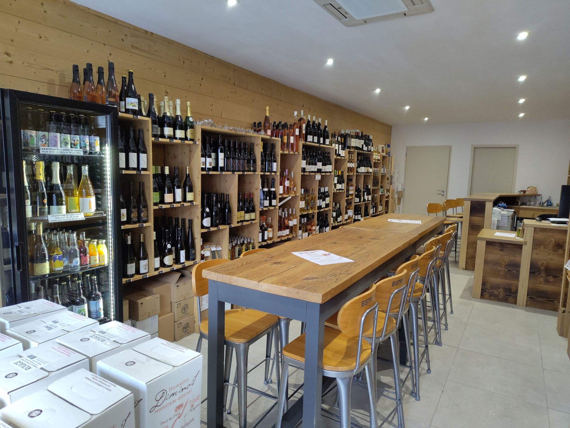 intérieur d'un magasin de caviste, des étagère pleine de bouteille en fond, une grande table pour soirée au milieu, ambiance chaleureuse typique de montagne donnée par le mur et le bar en bois