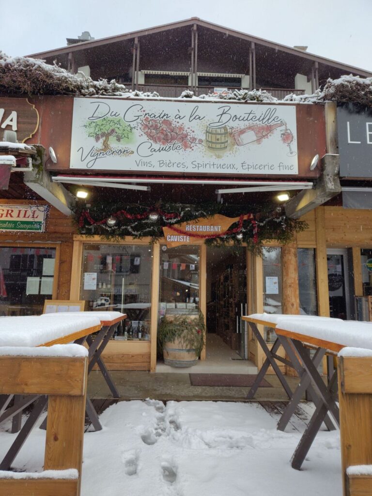 Façade extérieur d'un magasin de caviste, sous la neige, des tables de bois au premier plan grande baie vitrée du magasin en fond, pancarte du vigneron Du Grain à la Bouteille au dessus, ambiance typique de l'hiver en montagne.