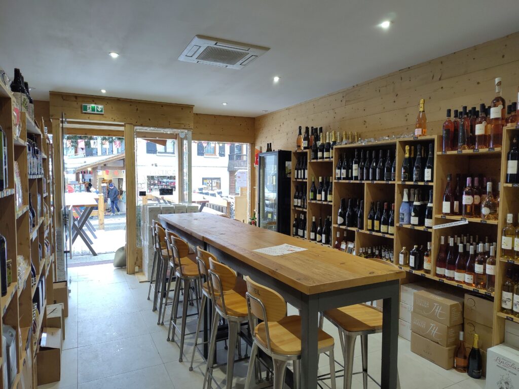intérieur d'un magasin de caviste, des étagères pleine de bouteille en fond, une grande table pour soirée au milieu, ambiance chaleureuse typique de montagne donnée par le mur et le bar en bois